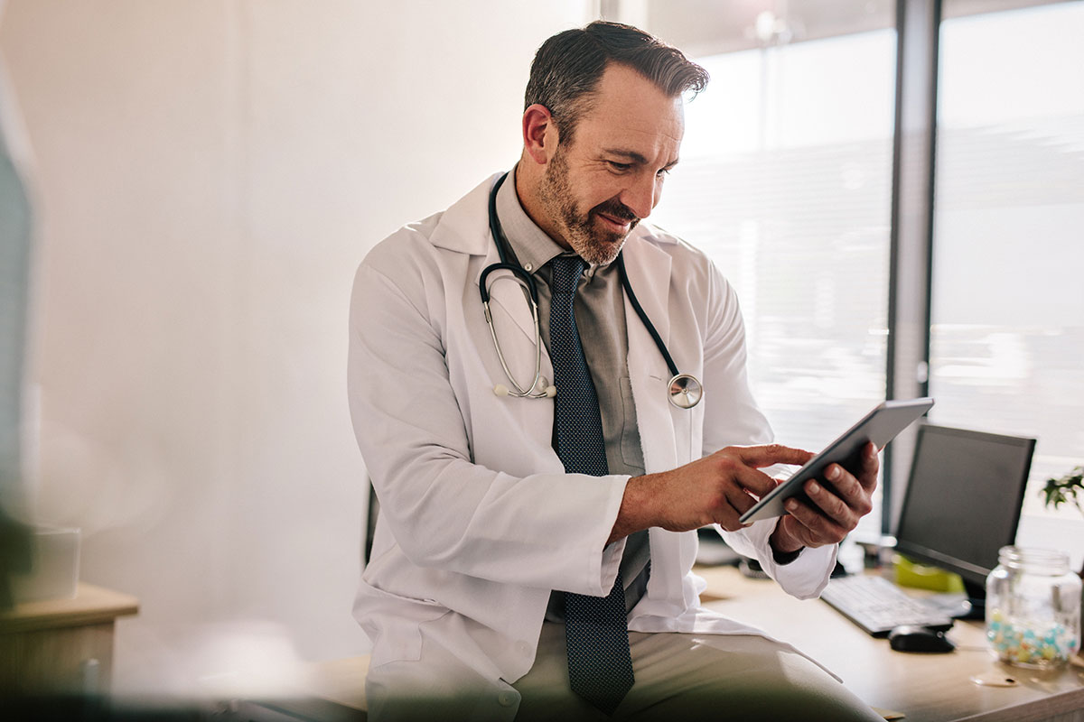 AdobeStock_249496776 A doctor reviews patient information on a tablet beside a laptop, illustrating efficiency and compliance in healthcare IT management.