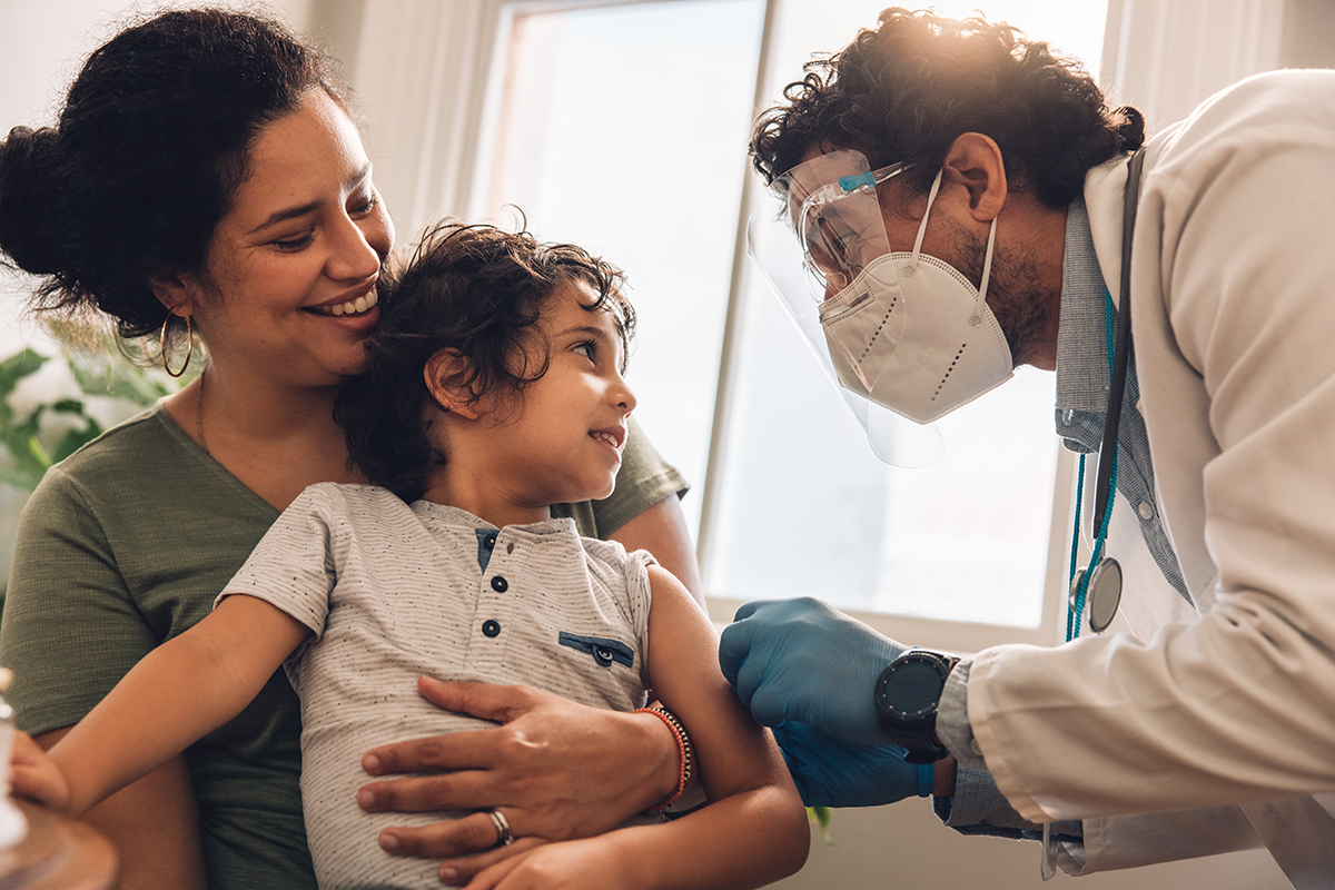 Vaccination during pandemic A smiling doctor wearing a mask greets a young child sitting on their mother’s lap, symbolizing compassionate and efficient healthcare supported by C2 ITSM.