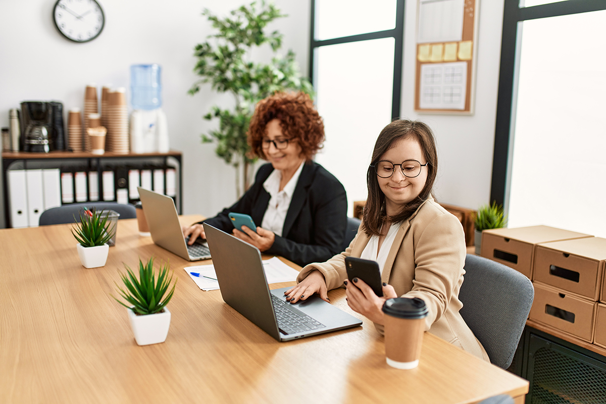 Group of two women working at the office. Mature woman and down syndrome girl working at inclusive teamwork. Deux professionnelles administratives travaillant sur des portables dans un bureau organisé, analysant des demandes internes