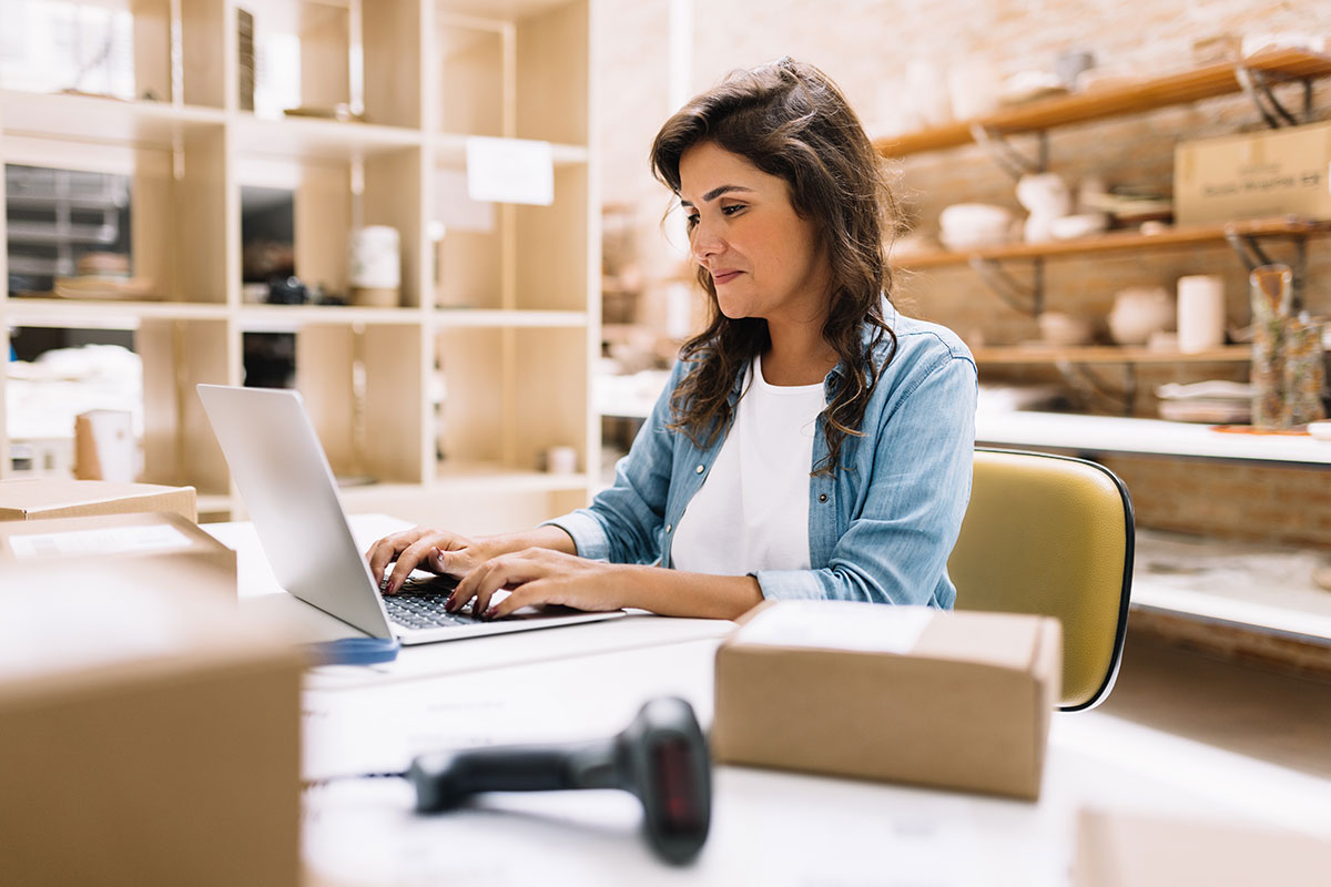 AdobeStock_605678244 Woman working on a laptop in a warehouse office, managing inventory and equipment data.