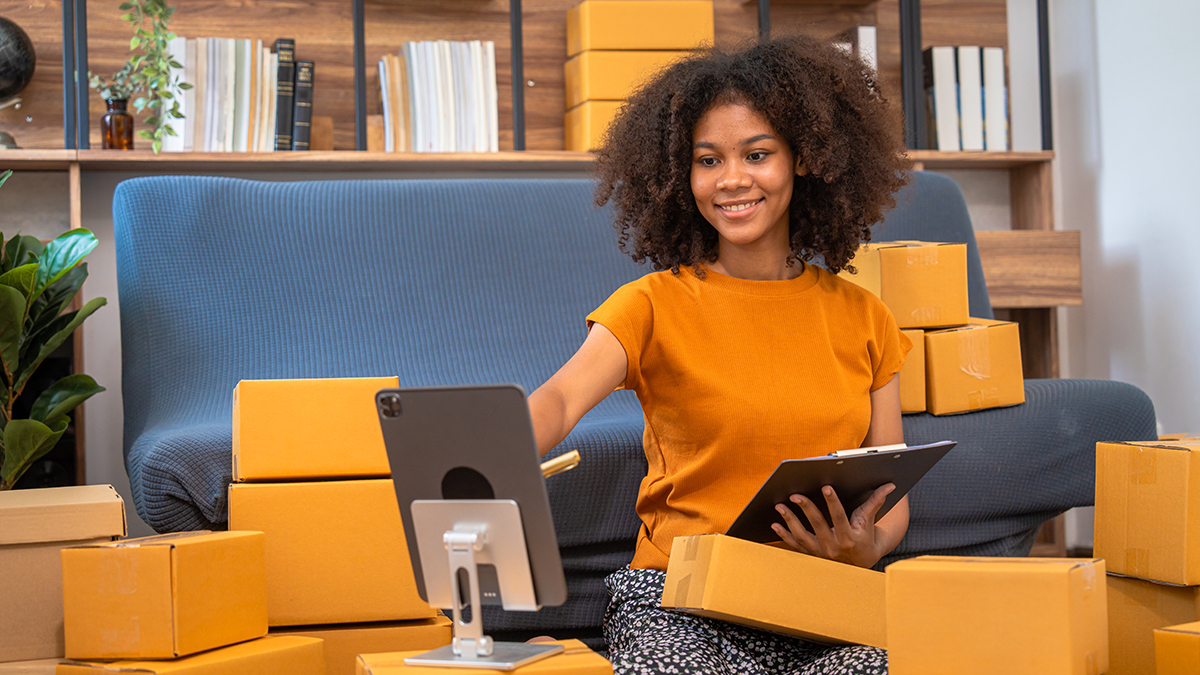 African American business woman working at warehouse preparing S Employée entourée de boîtes de livraison, utilisant une tablette et un terminal numérique pour gérer les demandes de service.