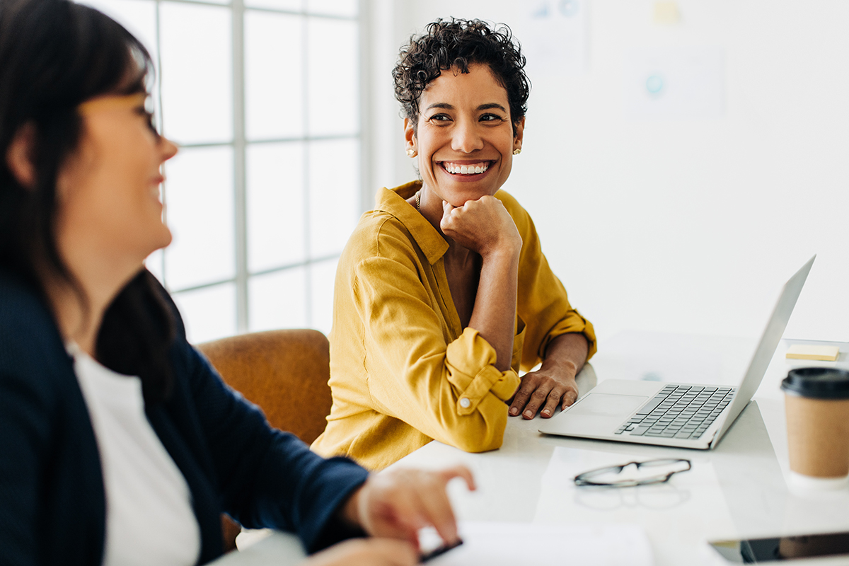 Happy business woman talking to her colleague in a meeting Two colleagues smiling and collaborating at a desk with a laptop, representing structured ITIL-inspired change management.